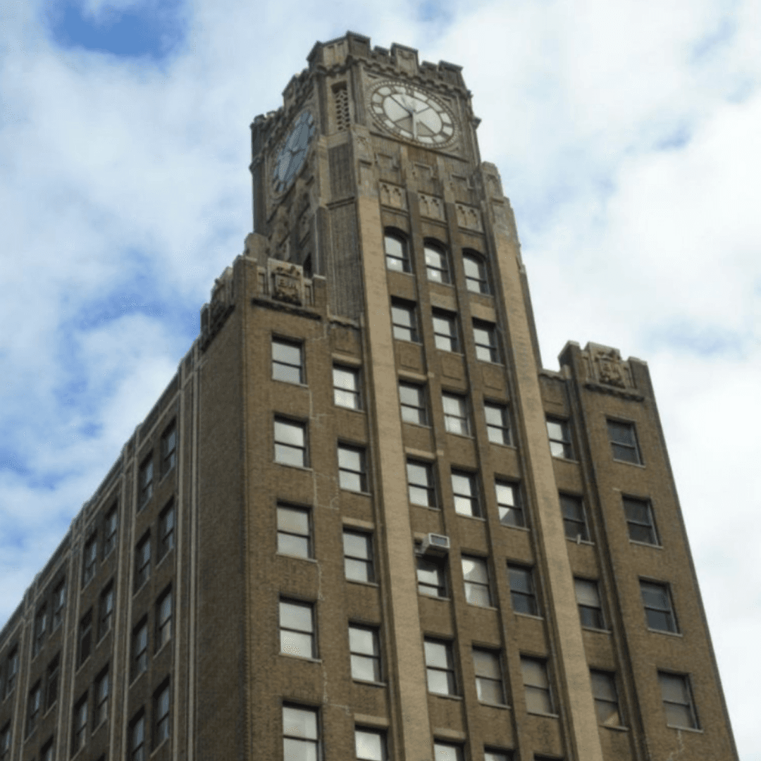 Street level view of the LIC Clock Tower with a cloudy blue sky behind it.